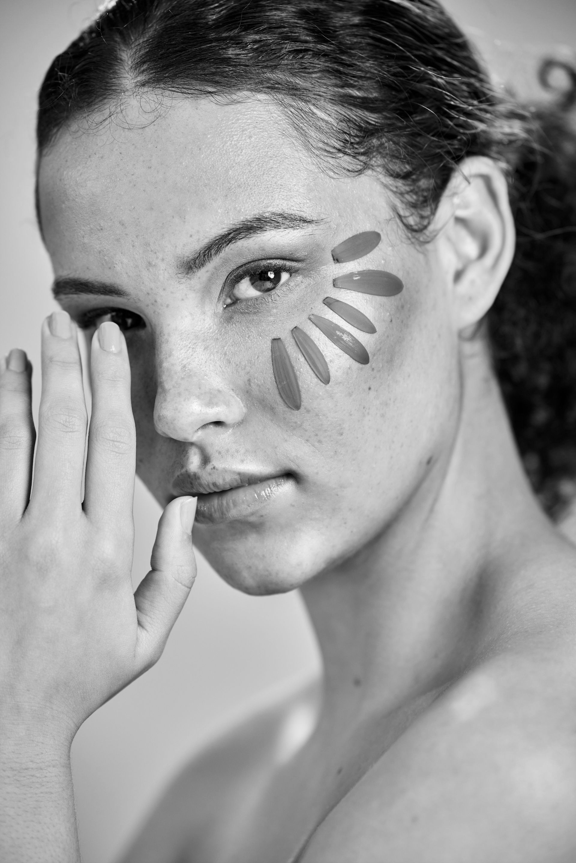 Une personne pose avec des marques de maquillage en forme de pétales déployées sur sa pommette, la main tenue près de son visage.