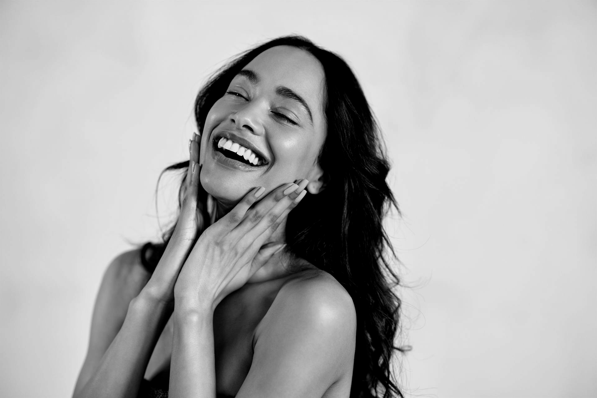 Une personne souriante aux cheveux longs pose ses mains sur ses joues, l'air heureux sur un fond uni.