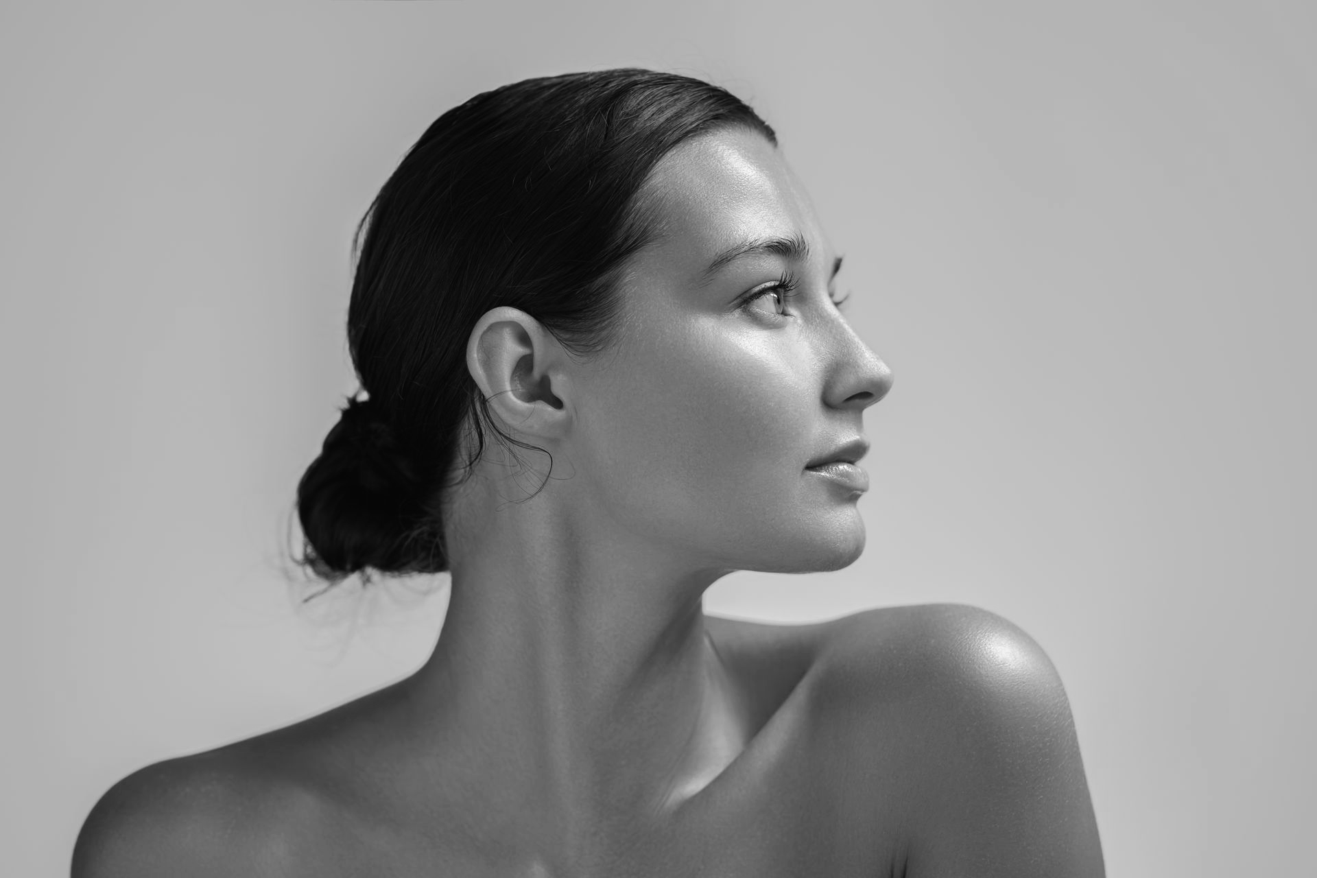 Portrait en studio, en noir et blanc, de profil, d'une personne aux cheveux coiffés en chignon bas, sur fond neutre.