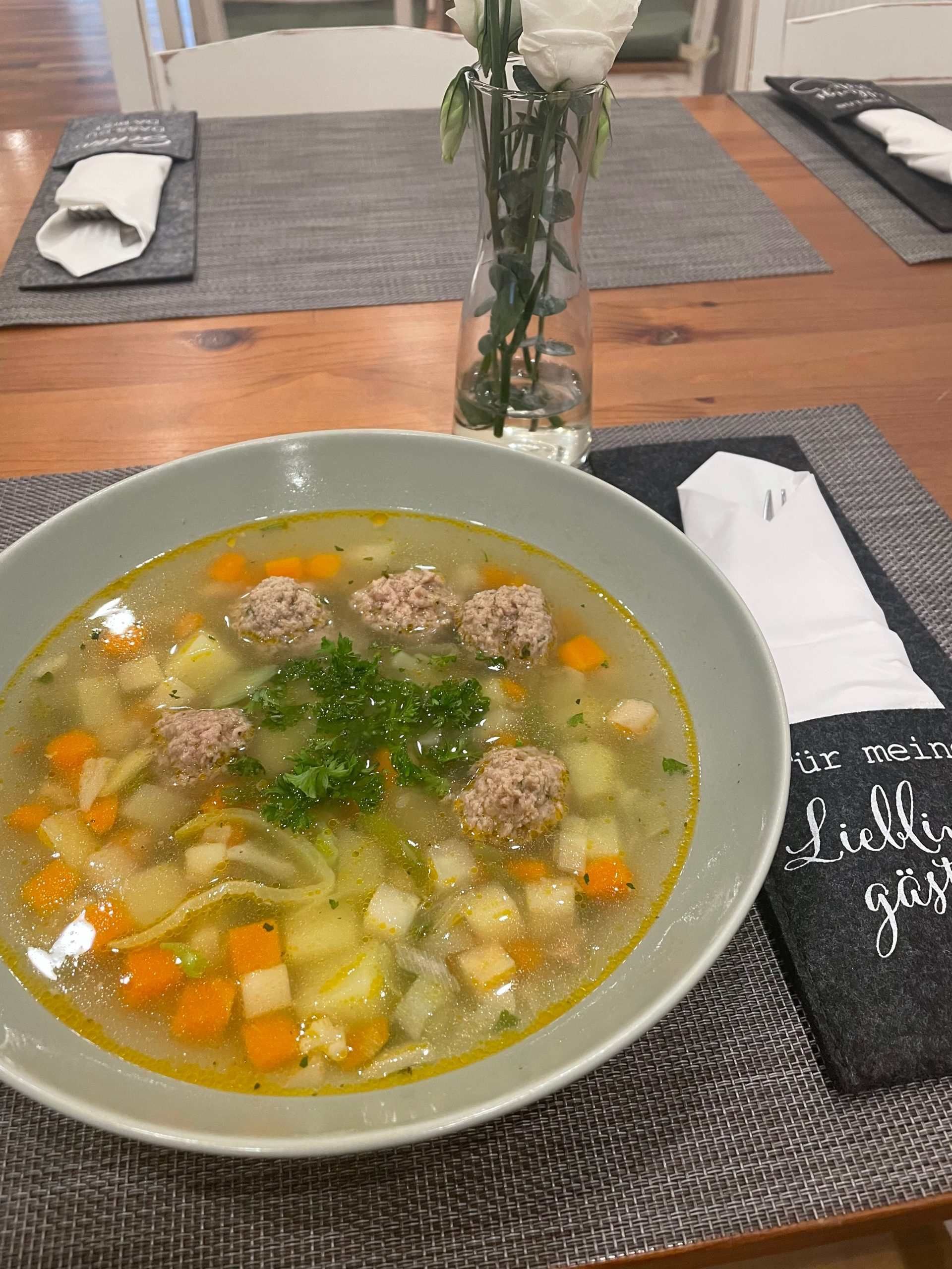 Auf einem Tisch steht eine Schüssel Suppe neben einer Vase mit Blumen. Mom & Son