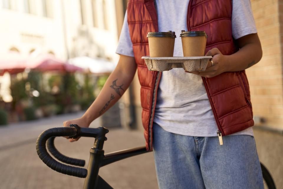 Ein Mann hält ein Tablett mit Kaffeetassen und steht neben einem Fahrrad. Mom & Son