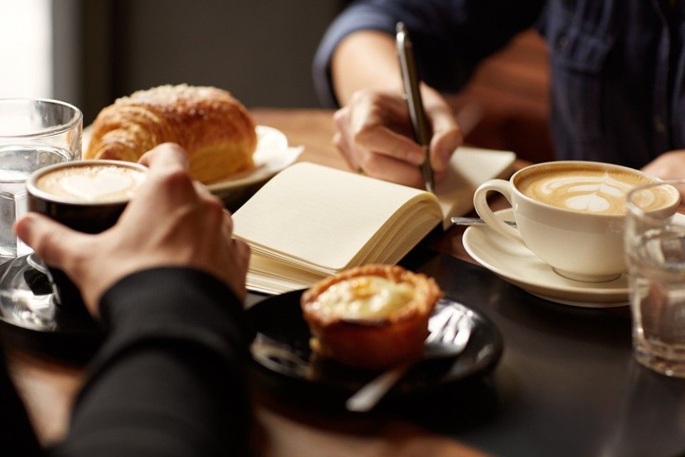 Eine Person schreibt in ein Notizbuch, während sie bei Kaffee und Gebäck an einem Tisch sitzt.