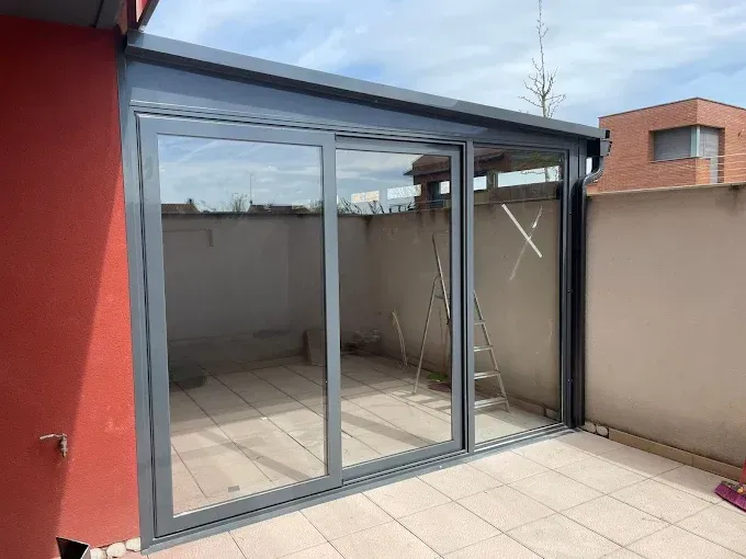 Patio cerrado con vidrio, puertas corredizas, marco gris, pared roja, piso de baldosas neutrales y paredes exteriores bajo un cielo despejado.