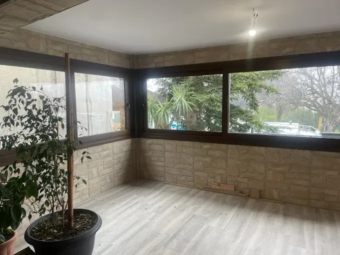 Terraza acristalada con paredes de azulejos, grandes ventanales con vistas a una calle arbolada y una gran planta en maceta.