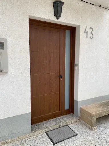 Puerta marrón con ventana lateral, número 43 arriba. Banco gris a la derecha, felpudo abajo.