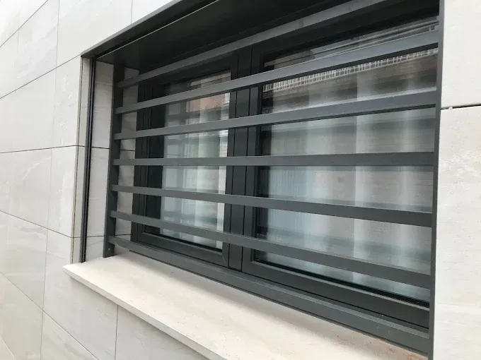 Ventana gris con rejas horizontales, dentro de una pared de azulejos blancos, con alféizar blanco.