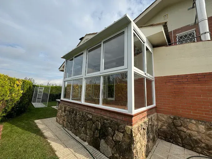 Terraza acristalada con marcos blancos y ventanas de cristal adosada a una casa de ladrillo y piedra.