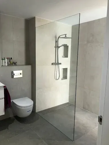 Baño moderno con azulejos grises, ducha de cristal, inodoro montado en la pared y accesorios cromados.