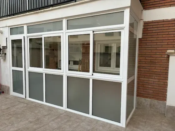Terraza acristalada con marcos blancos, ventanas de cristal y paneles inferiores esmerilados, adosada a un edificio de ladrillo.