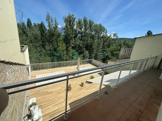 Balcón con barandilla de cristal, terraza de madera y árboles contra un cielo azul.