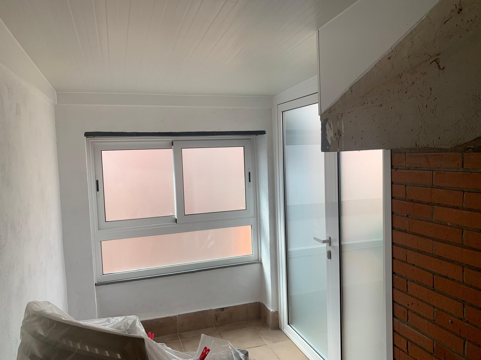 Vista interior de una habitación con ventana, puerta y pared de ladrillo; paredes y techo blancos.