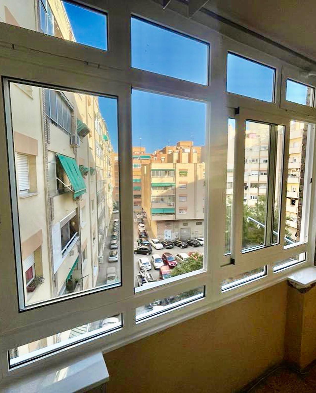 Vista desde un balcón cerrado. A través de las ventanas con marcos blancos se ve una calle con edificios y coches.
