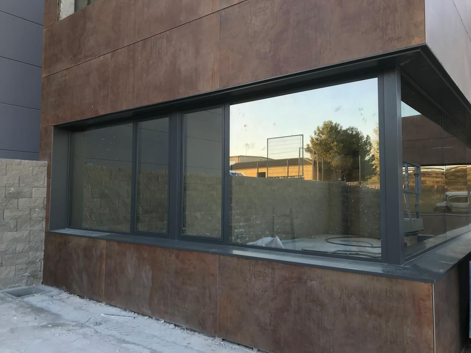 Gran ventana rectangular en la fachada marrón de un edificio, que refleja una obra en construcción y el cielo.