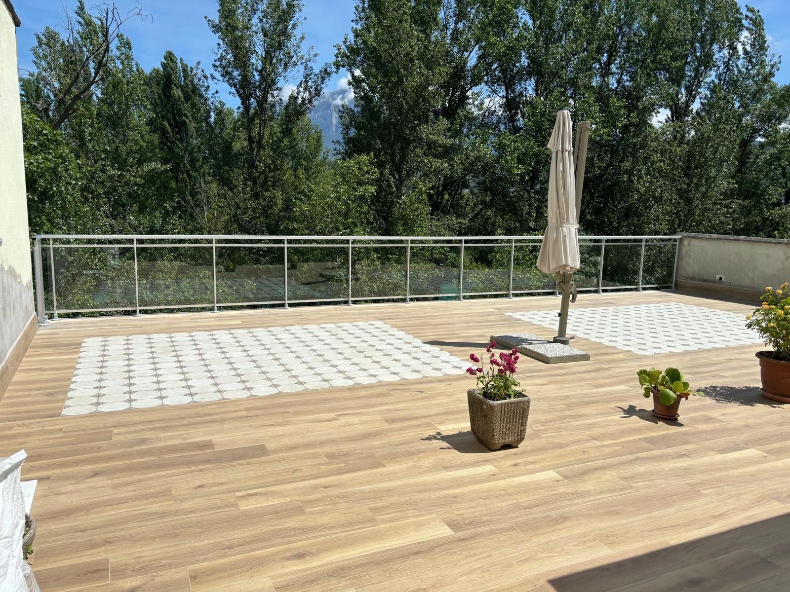 Patio soleado en la azotea con terraza de madera, adoquines de piedra, barandilla de vidrio y sombrilla; árboles al fondo.