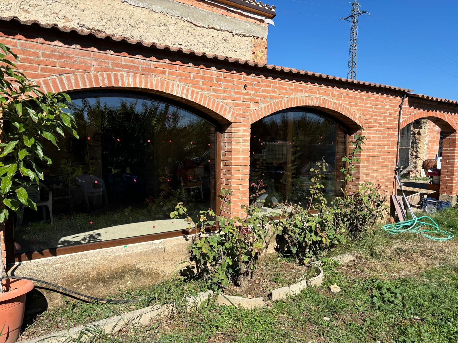 Edificio de ladrillo con ventanas arqueadas, que refleja una escena exterior soleada con algo de vegetación.