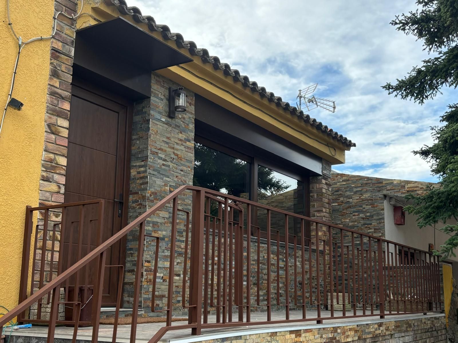 Casa con fachada de piedra, con puerta, barandilla y molduras de ventanas de color marrón, bajo un cielo nublado.