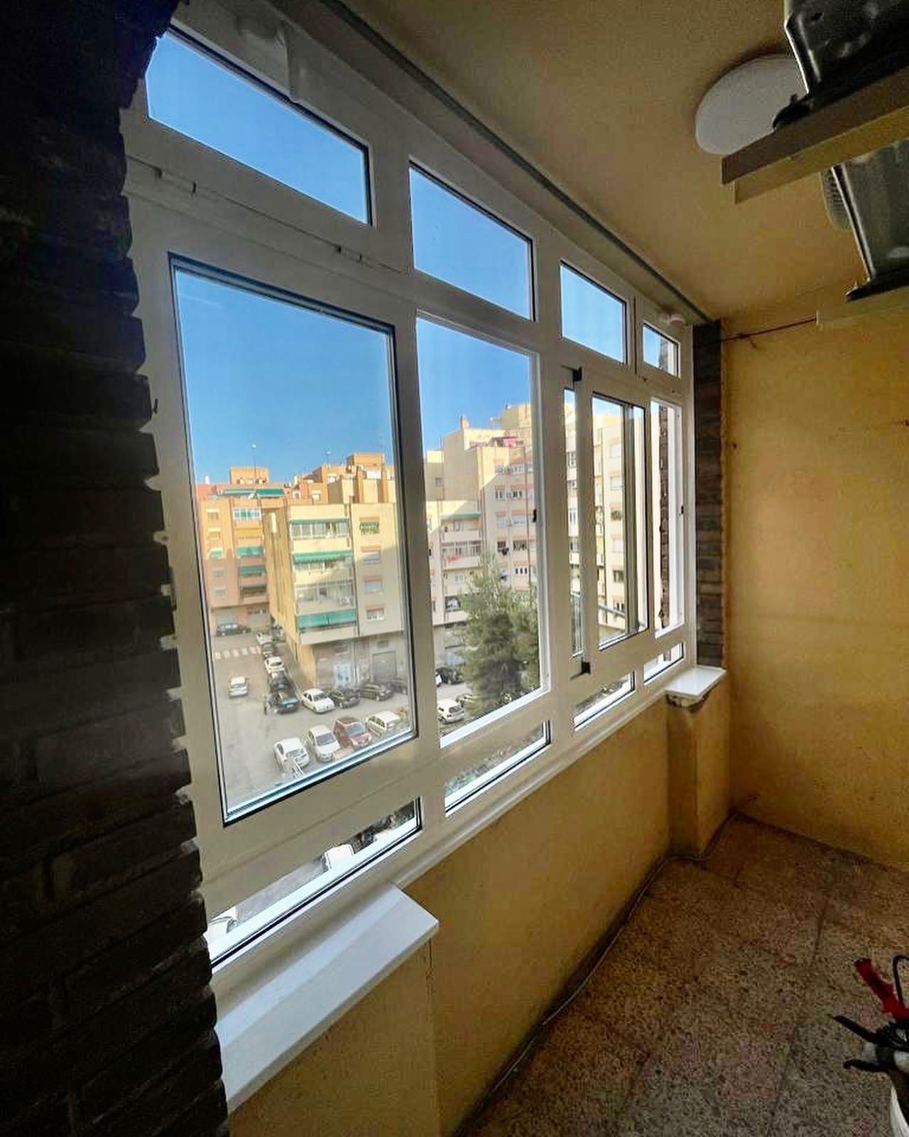 Vista de una calle de la ciudad desde el interior de una ventana de balcón. La ventana tiene un marco blanco y las paredes son beige.
