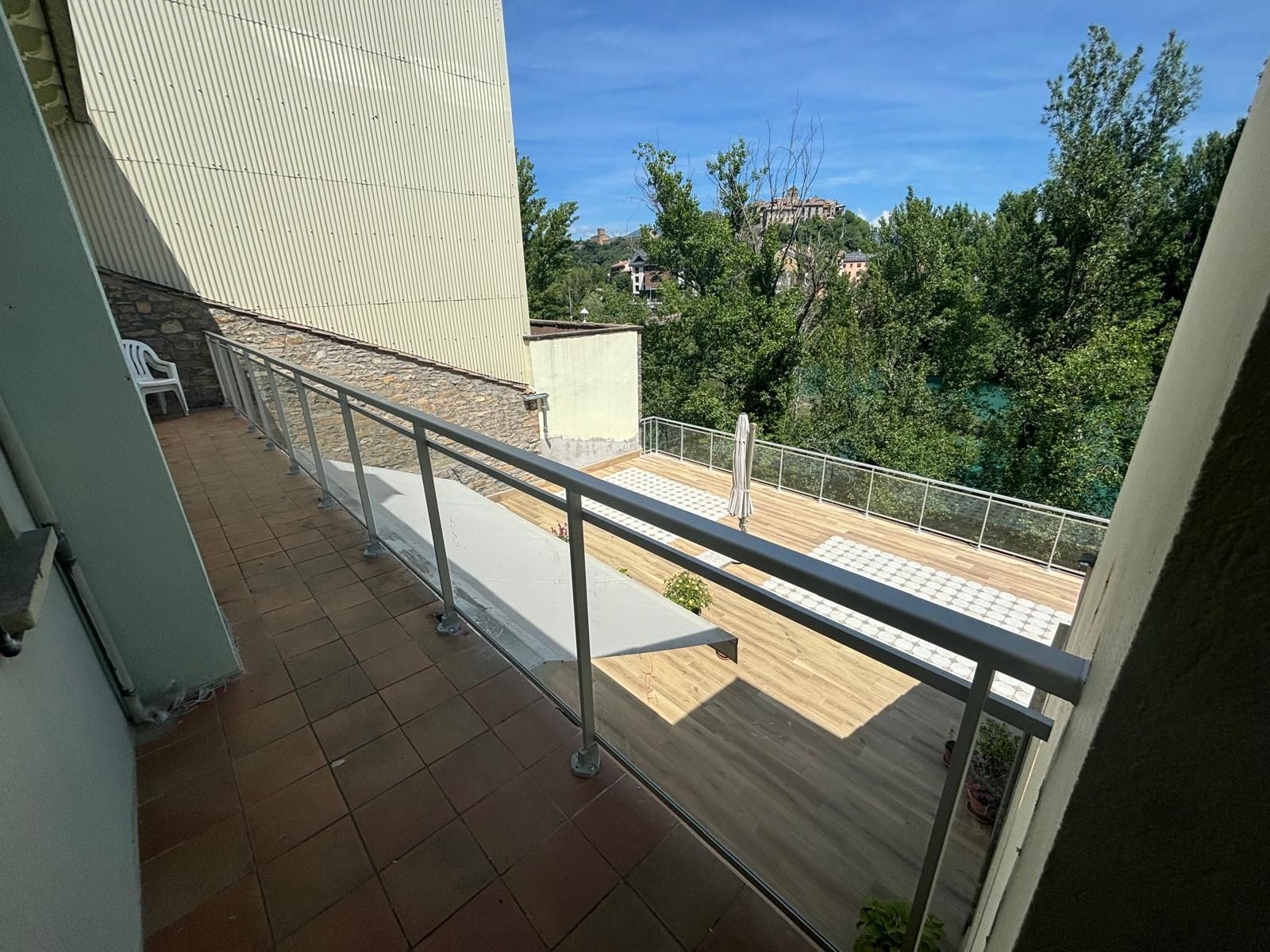 Balcón con barandillas de vidrio, madera de color claro, piso de baldosas, con vista a los árboles y al cielo azul.