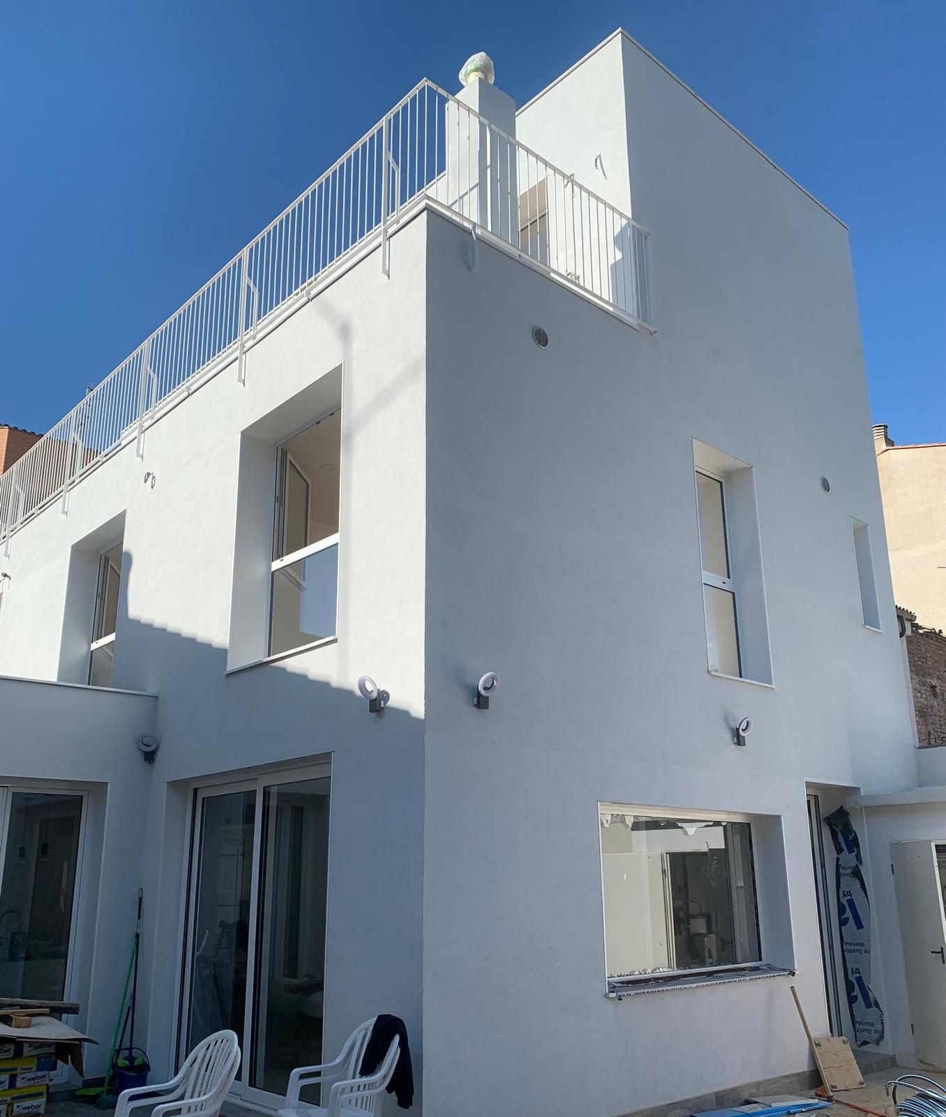 Edificio blanco de dos plantas con balcón en la azotea; cielo azul claro. Ventanas visibles.