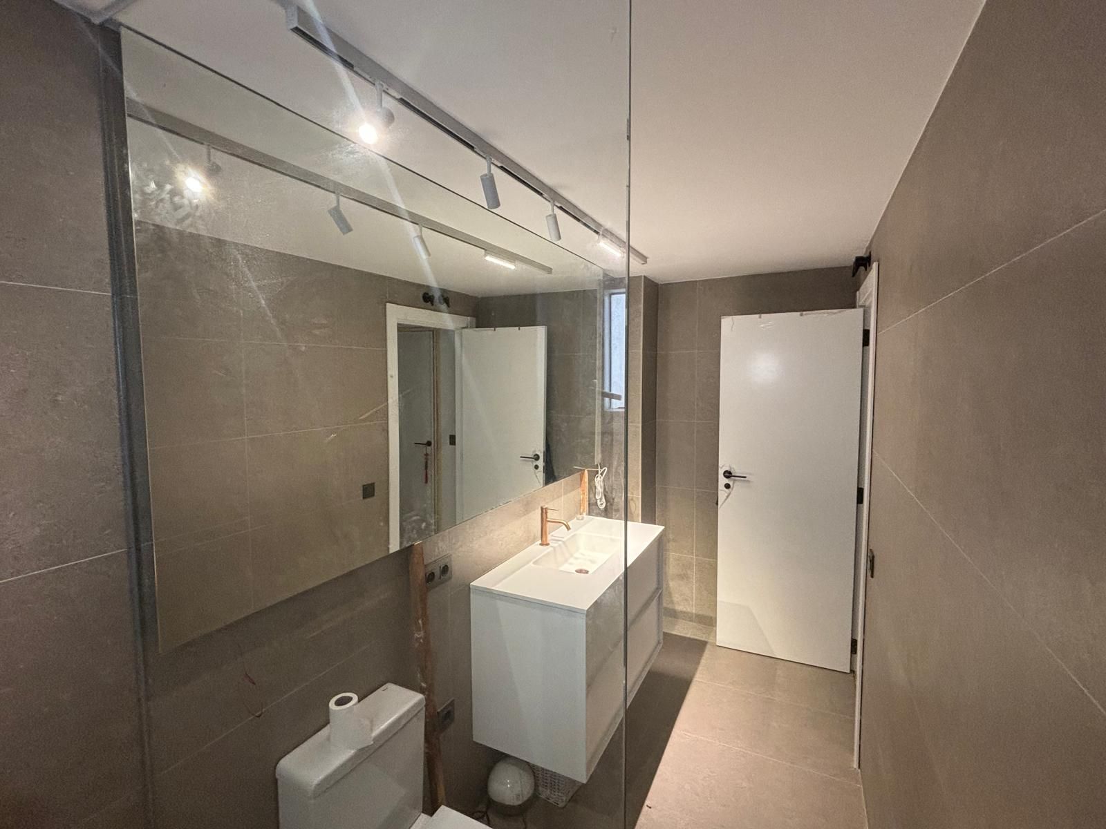 Baño moderno con espejo grande, lavabo e inodoro blancos. Paredes de azulejos grises y puerta blanca.