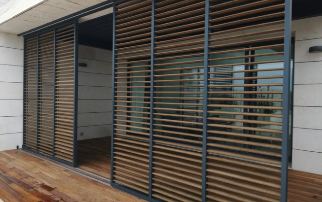 Mamparas corredizas de listones de madera sobre una terraza, que cubren parcialmente una puerta de vidrio que da a una habitación, con un piso de madera clara.