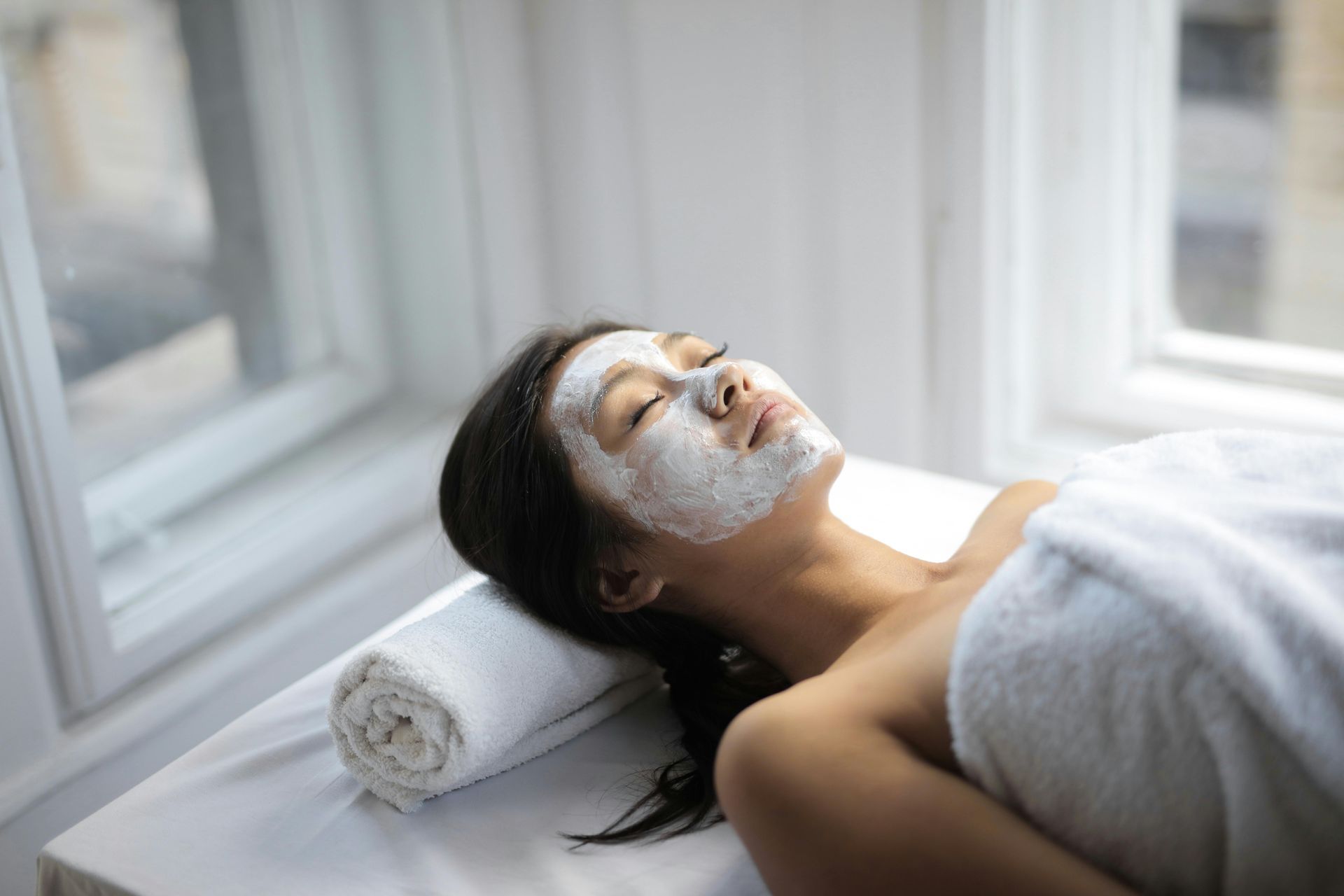 Mujer con mascarilla facial relajándose en una mesa de spa cerca de una ventana.