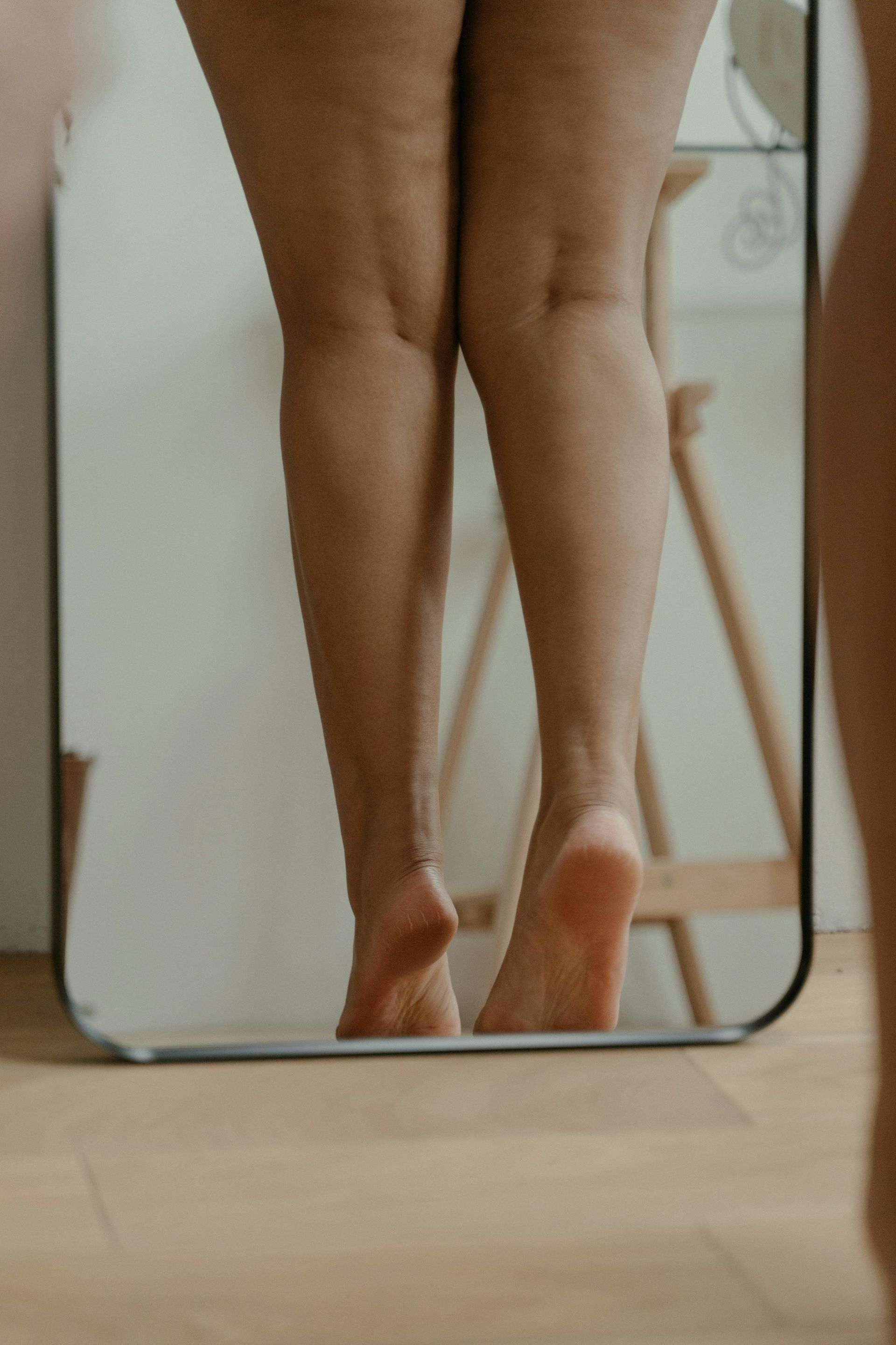 Piernas y pies de una persona parada de puntillas frente a un espejo, reflejando su forma.