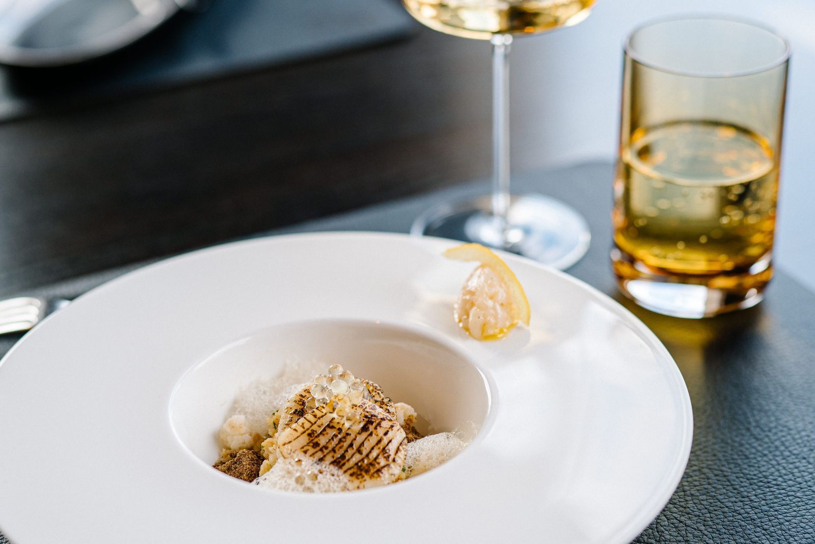 Ein Gourmetgericht mit Jakobsmuschel auf einem weißen Teller und ein Glas Wein im Restaurant 1831