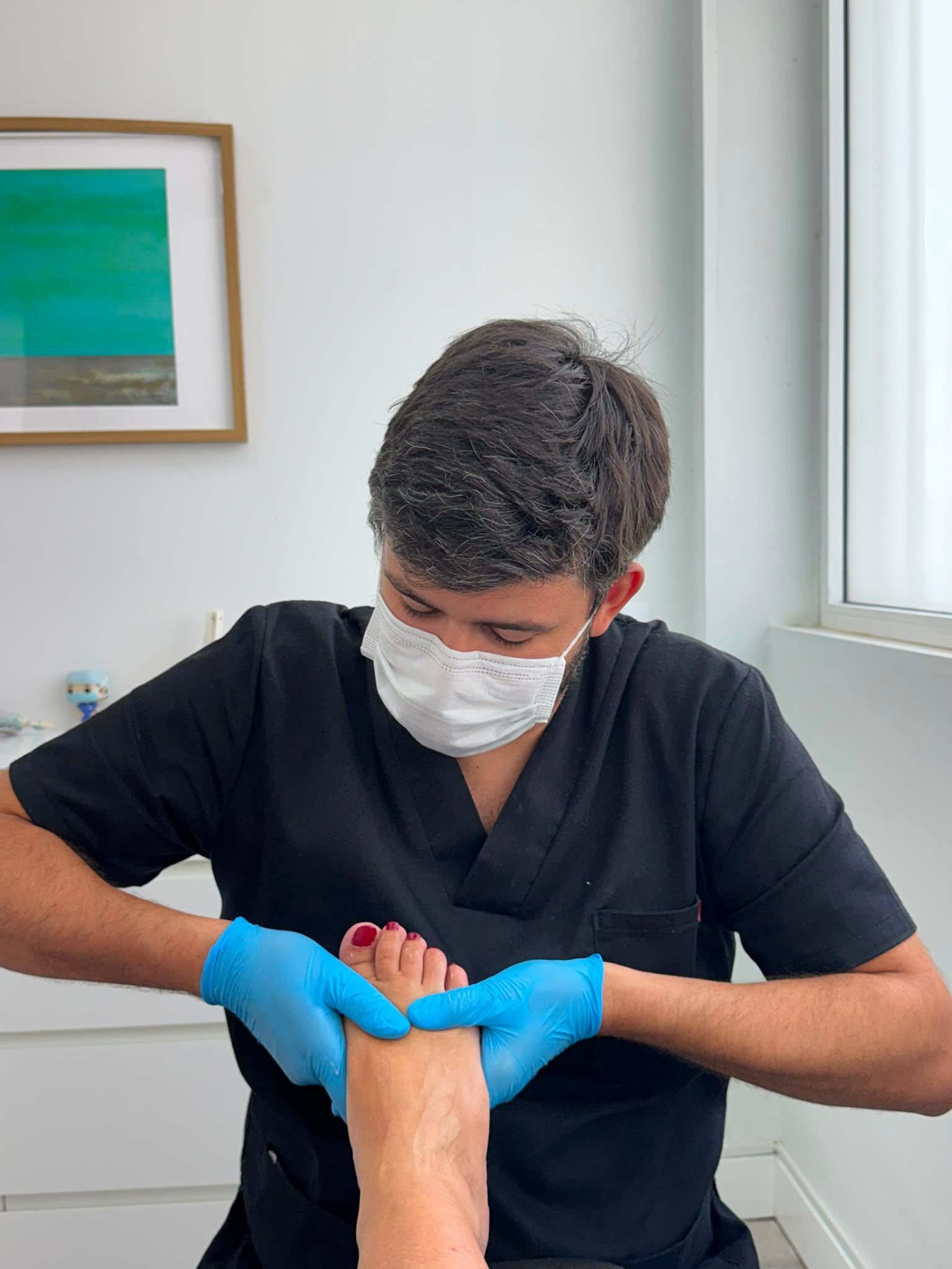 Un trabajador sanitario con bata negra y mascarilla atiende la mano enguantada de azul de un paciente cerca de una ventana.