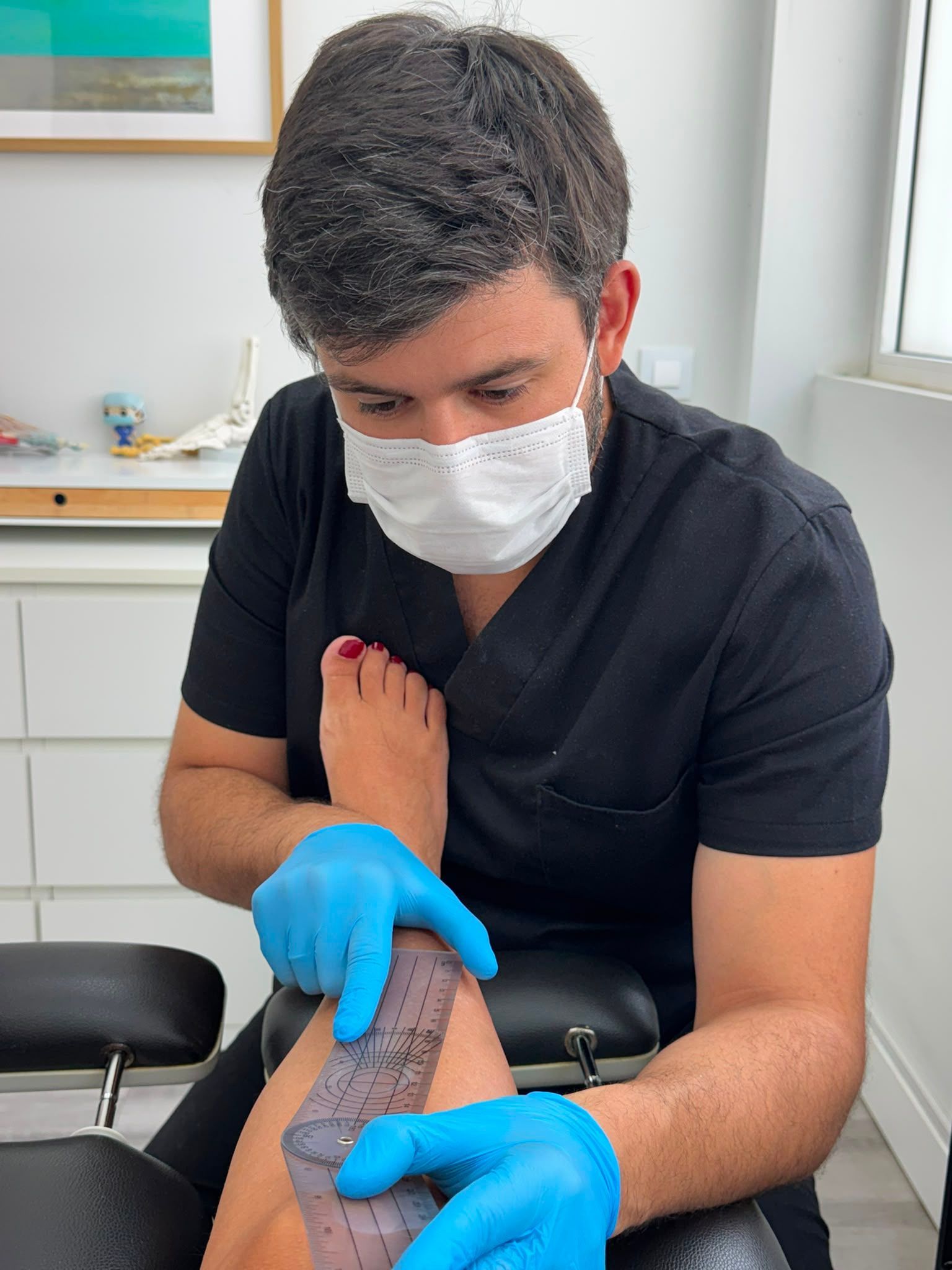 Un médico con mascarilla y guantes azules examina la pierna de un paciente en una sala de exploración médica.