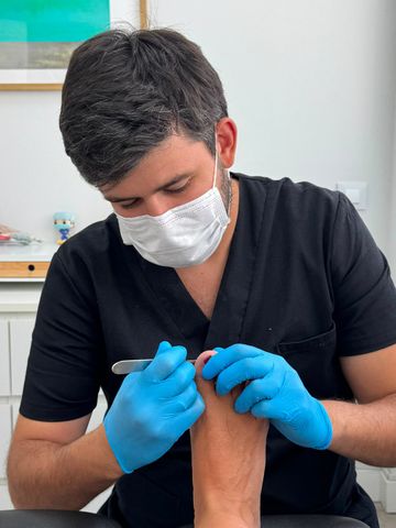 Podólogo con bata negra y guantes azules examinando el pie de un paciente en un consultorio médico.