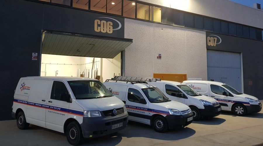Tres furgonetas de reparto blancas aparcadas frente a un muelle de carga de un almacén al anochecer.