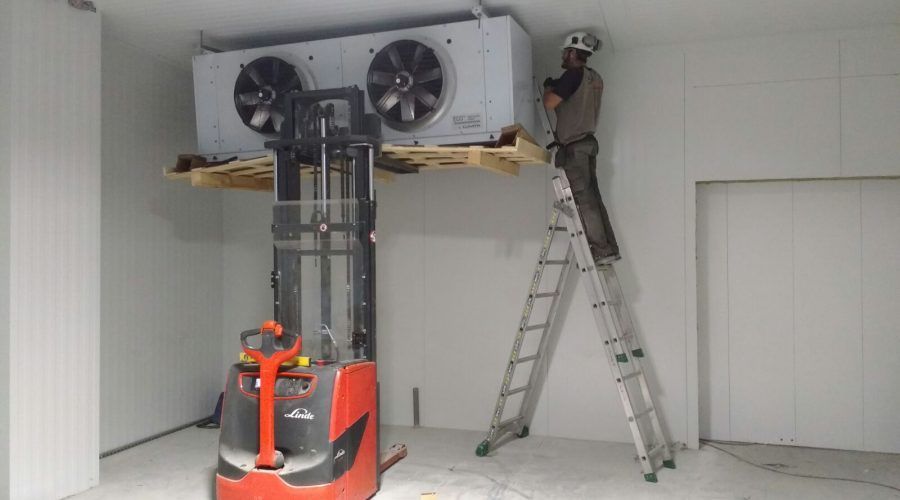 Trabajadores instalando una gran unidad de climatización en una habitación blanca utilizando escaleras y una carretilla elevadora.