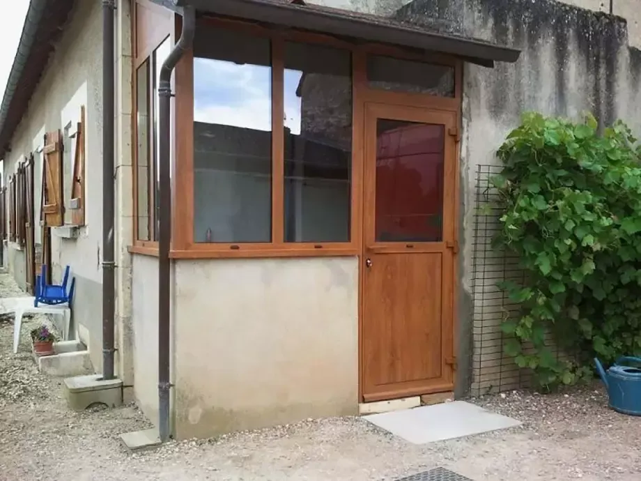 Porte d’entrée en bois et fenêtre sur la façade d’une petite maison beige, avec une plante en pot à côté de l’entrée.