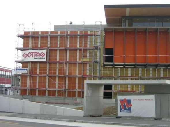 Ein mehrstöckiges, im Bau befindliches Gebäude mit orangefarbenen Verkleidungen, Gerüsten und Baustellenbannern mit Firmenlogo.