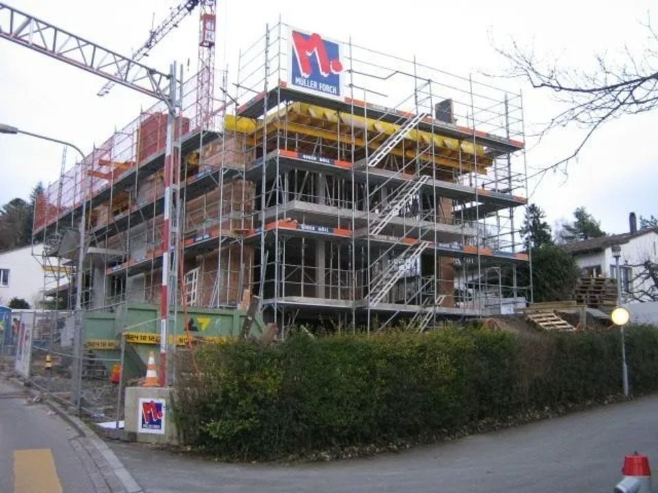 Mehrstöckiges, im Bau befindliches Gebäude, umgeben von einem Gerüst, darüber ein Kran und im Vordergrund eine Hecke.