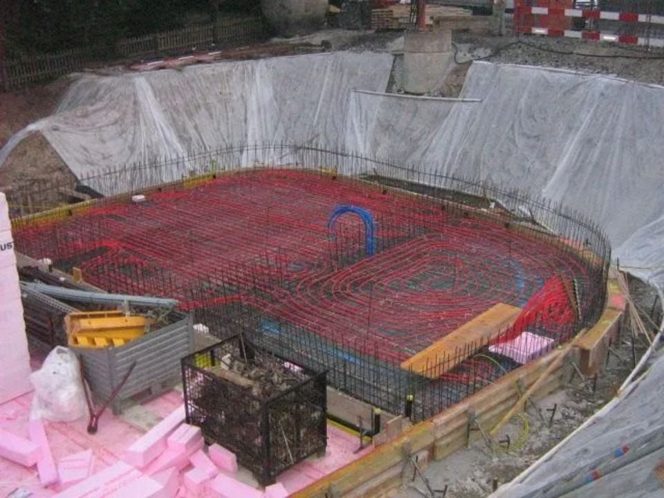Auf einer Baustelle ist ein Fundament mit rotem Metallarmierungsgewebe und Holzschalung in einer ausgehobenen Baugrube zu sehen.