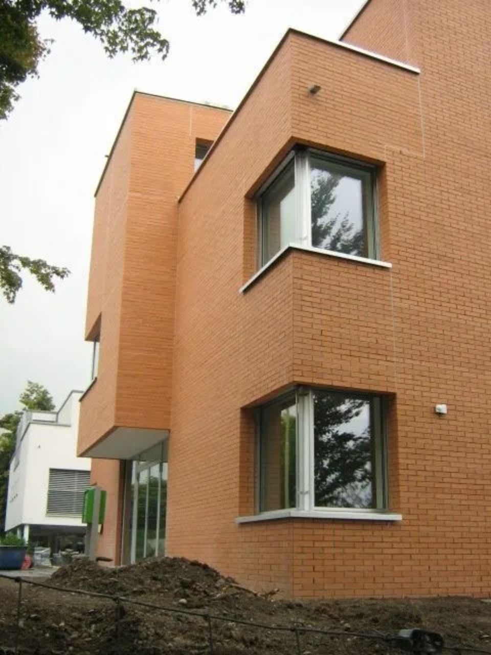 Ein modernes, zweistöckiges Gebäude aus orangefarbenen Ziegeln mit Eckfenstern und ein angrenzendes weisses Gebäude unter bewölktem Himmel.