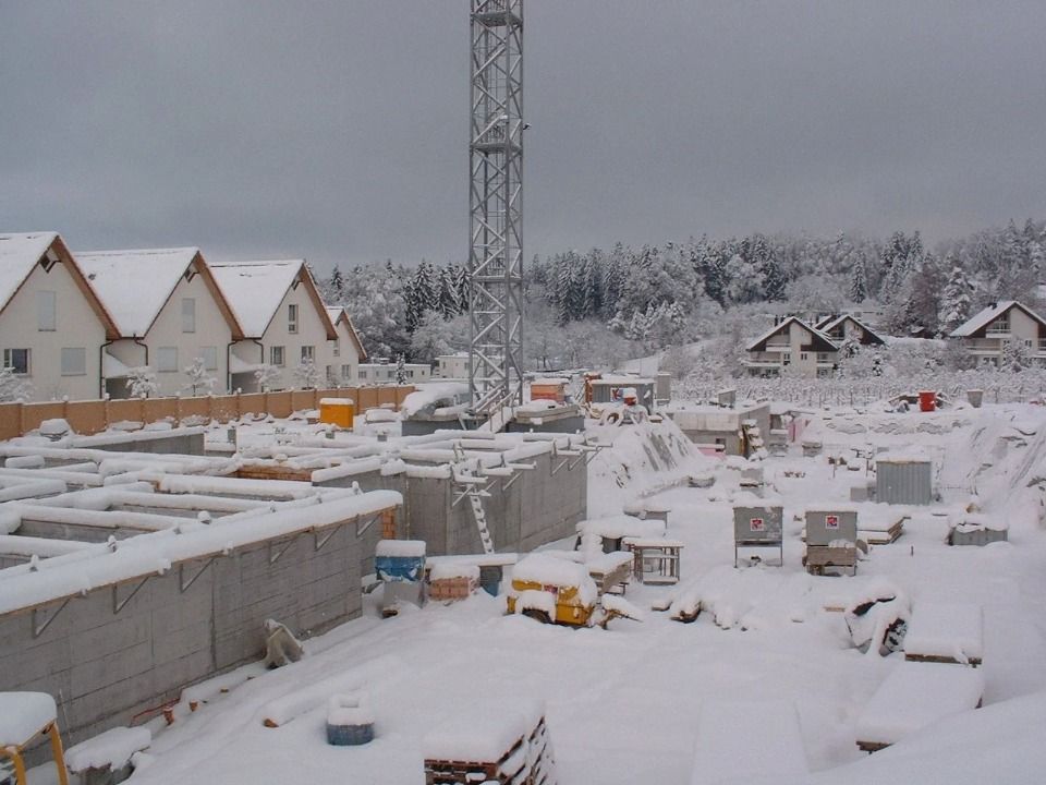 Eine verschneite Baustelle mit Betonfundamenten, einem hohen Turmdrehkran und im Hintergrund nahegelegenen Wohnhäusern.