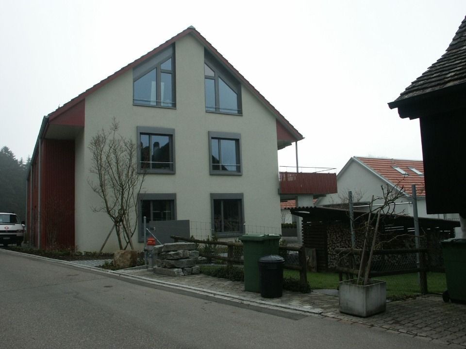 An einem bewölkten Tag steht neben einer Strasse ein modernes dreistöckiges Haus mit heller Fassade und dunkel gerahmten Fenstern.