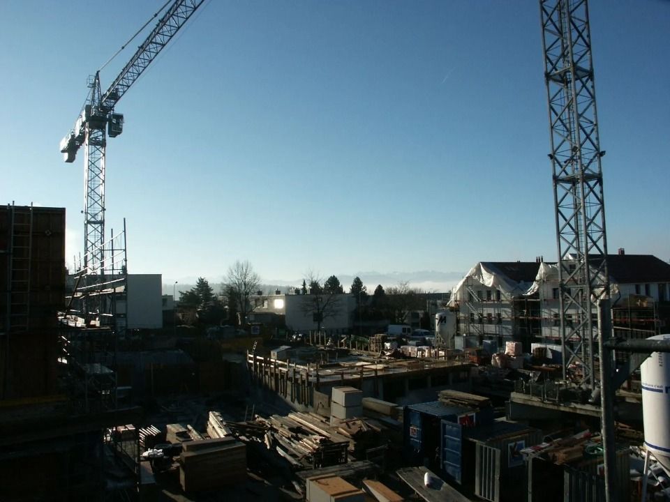 Eine Baustelle mit zwei grossen Turmdrehkranen, die ein unfertiges Gebäudefundament und Materialstapel überblicken.