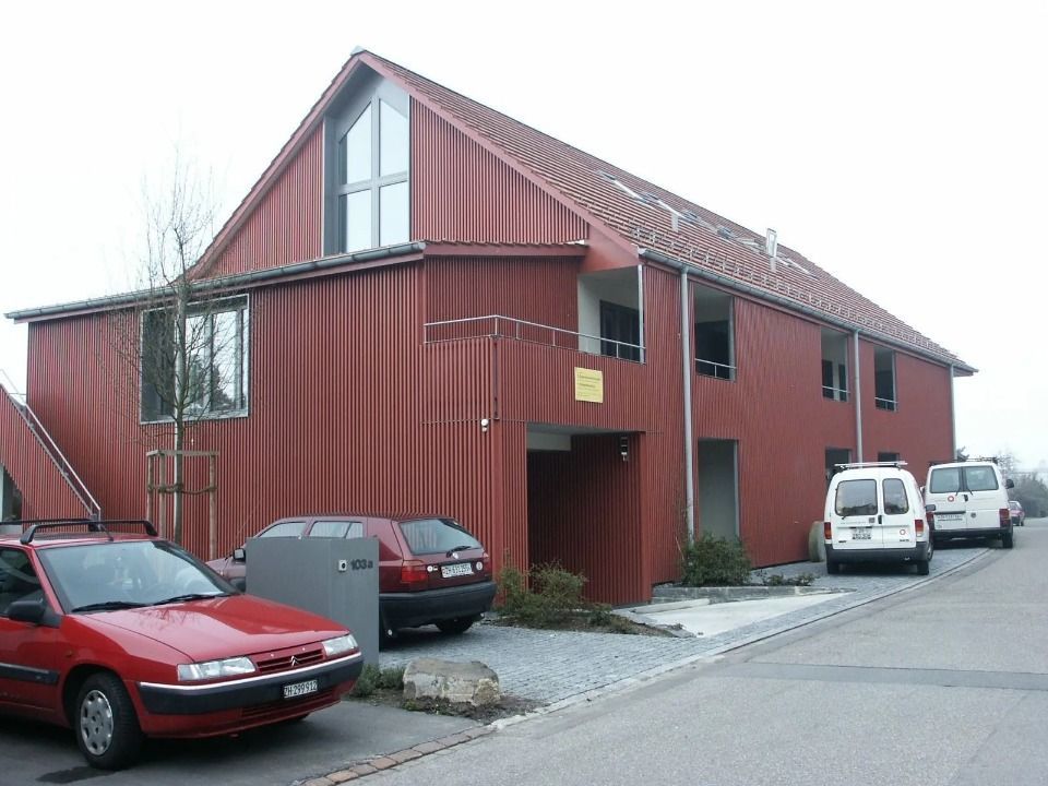 Ein zweistöckiges Gebäude mit rötlich-brauner Wellblechverkleidung, einem Satteldach und mehreren geparkten Autos im Vordergrund.