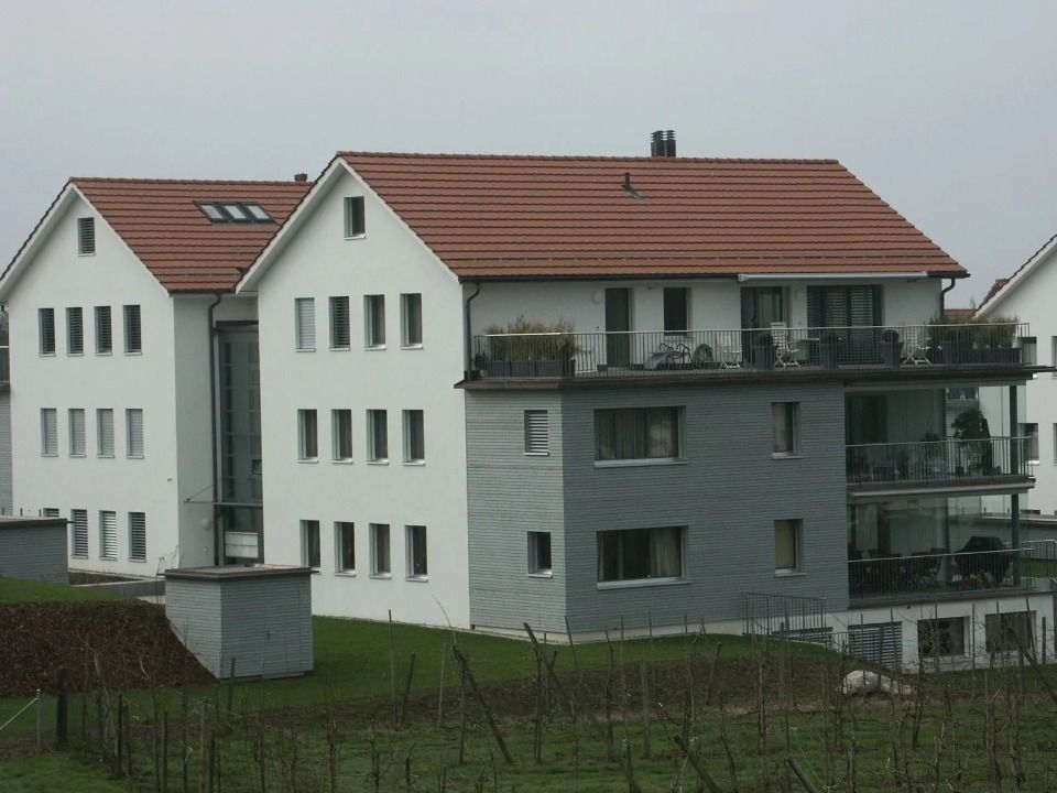 Zwei moderne, mehrstöckige Gebäude mit weissen Wänden, grauer Fassade und Terrakotta-Ziegeldächern vor einem bewölkten Himmel.