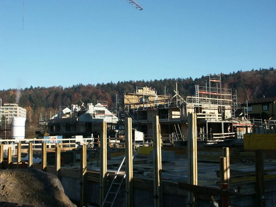 Eine Baustelle mit im Bau befindlichen Holzrahmenbauten, Gerüsten und Betonfundamentpfeilern unter blauem Himmel.