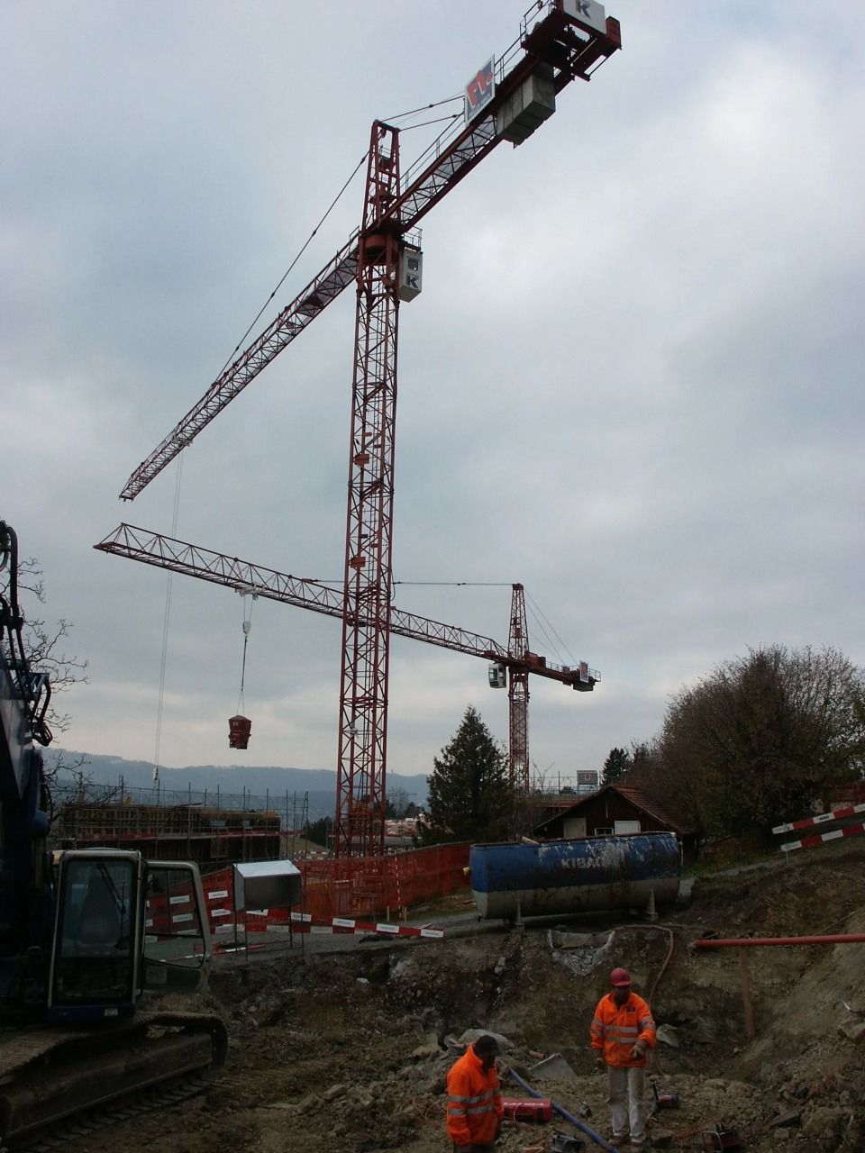 Zwei Bauarbeiter in orangefarbenen Westen auf einer Baustelle mit zwei hohen roten Kränen unter bewölktem Himmel.