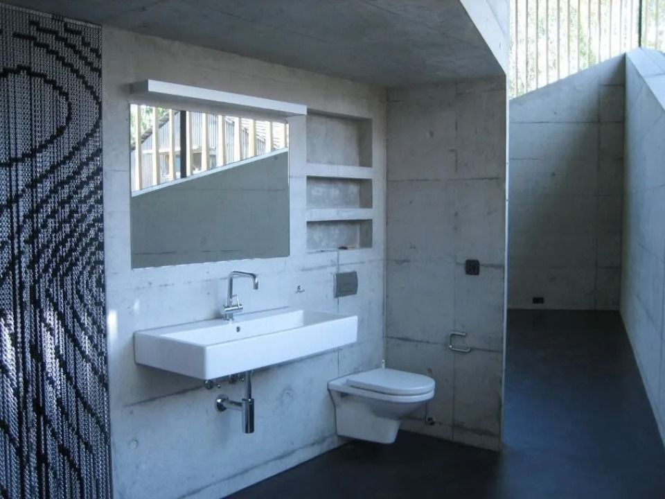 Modernes Badezimmer mit rohen Betonwänden, einem weissen, wandhängenden Waschbecken und WC, Wandregalen und einem gemusterten Perlenvorhang.