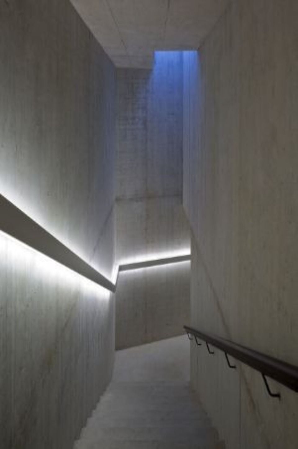 Minimalistisches Treppenhaus mit strukturierten Betonwänden, LED-Streifenbeleuchtung und einem dunklen Handlauf, der zu einer blau beleuchteten Öffnung führt.