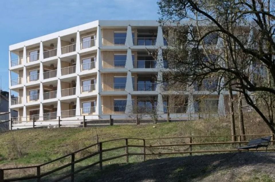 Ein modernes, vierstöckiges Wohnhaus mit gitterartigem Betonrahmen und Balkonen, gelegen auf einem grasbewachsenen Hügel.