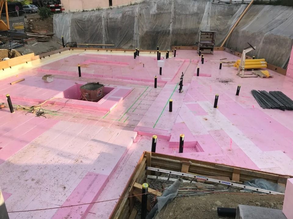 Baustelle mit einer Fundamentplatte, die mit rosafarbenen Dämmplatten bedeckt ist, aus der Rohre und Bewehrungsstahl herausragen.
