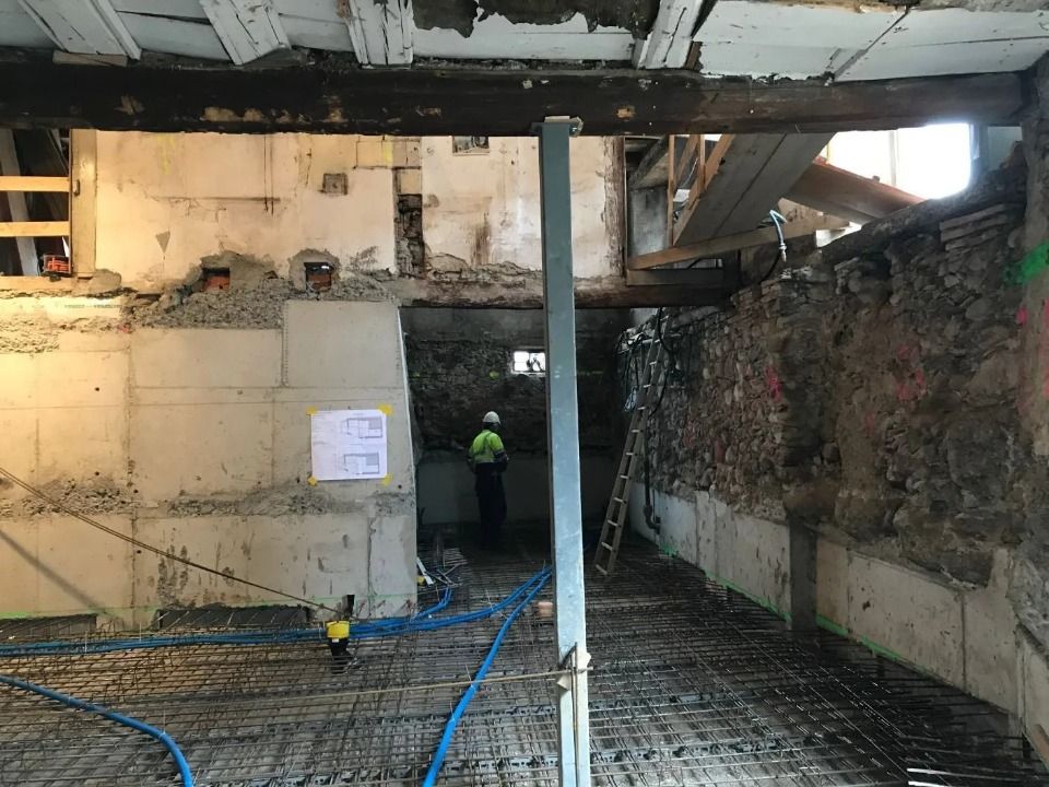 Auf einer Baustelle sind ein Arbeiter, ein Metallträger und freiliegende Steinmauern mit einer vorbereiteten Betonbodenfläche zu sehen.